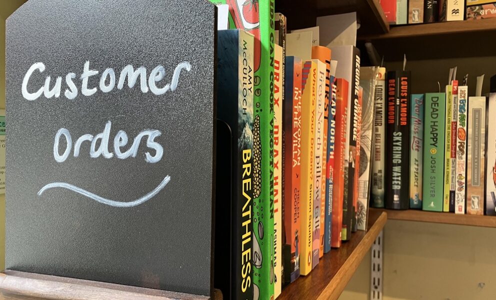A shelf of books with a chalked sign in the foreground reading "Customer Orders"